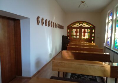 Wnętrze kaplicy. Wąskie pomieszczenie z rzędem drewnianych ławek. Z tyłu drewniane drzwi z witrażem.