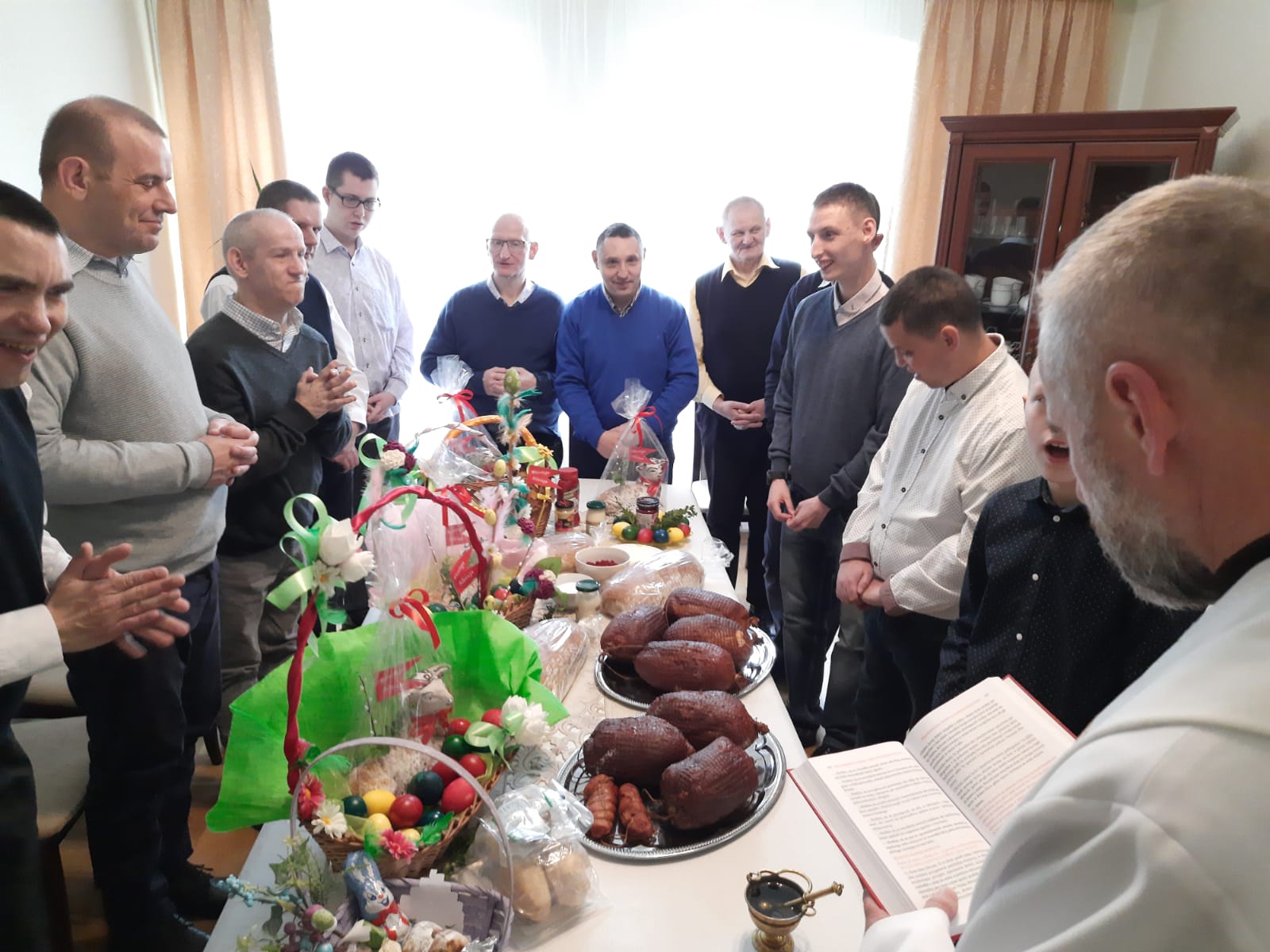 Zdjęcie wykonane w pokoju gościnnym podczas święcenia pokarmów przez księdza proboszcza. Przy obficie zastawionym stole zgromadzeni są mieszkańcy domu.