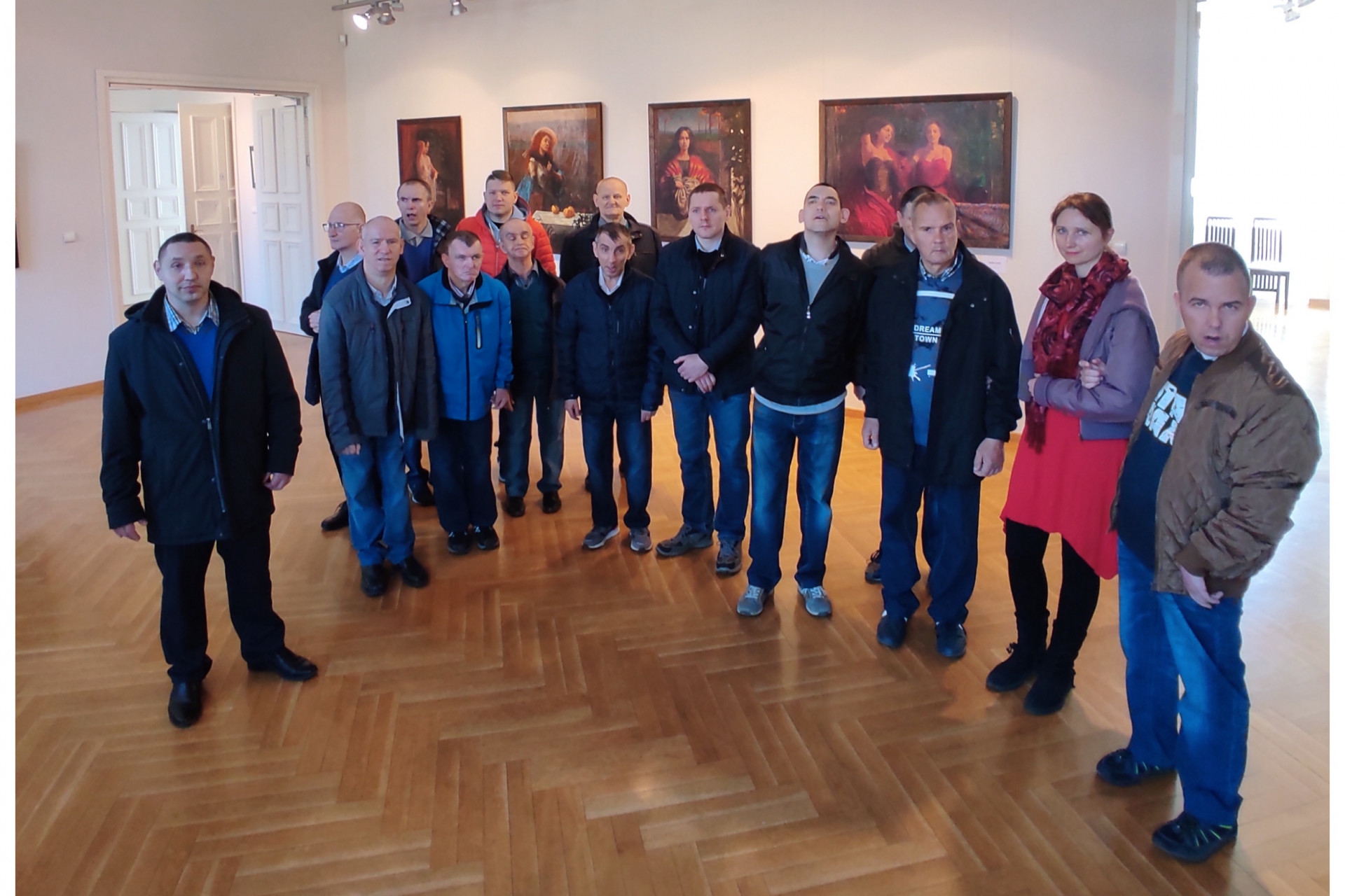 Zdjęcie grupowe podopiecznych wraz z terapeutami w galerii na tle monumentalnych obrazów stacji Drogi Krzyżowej.