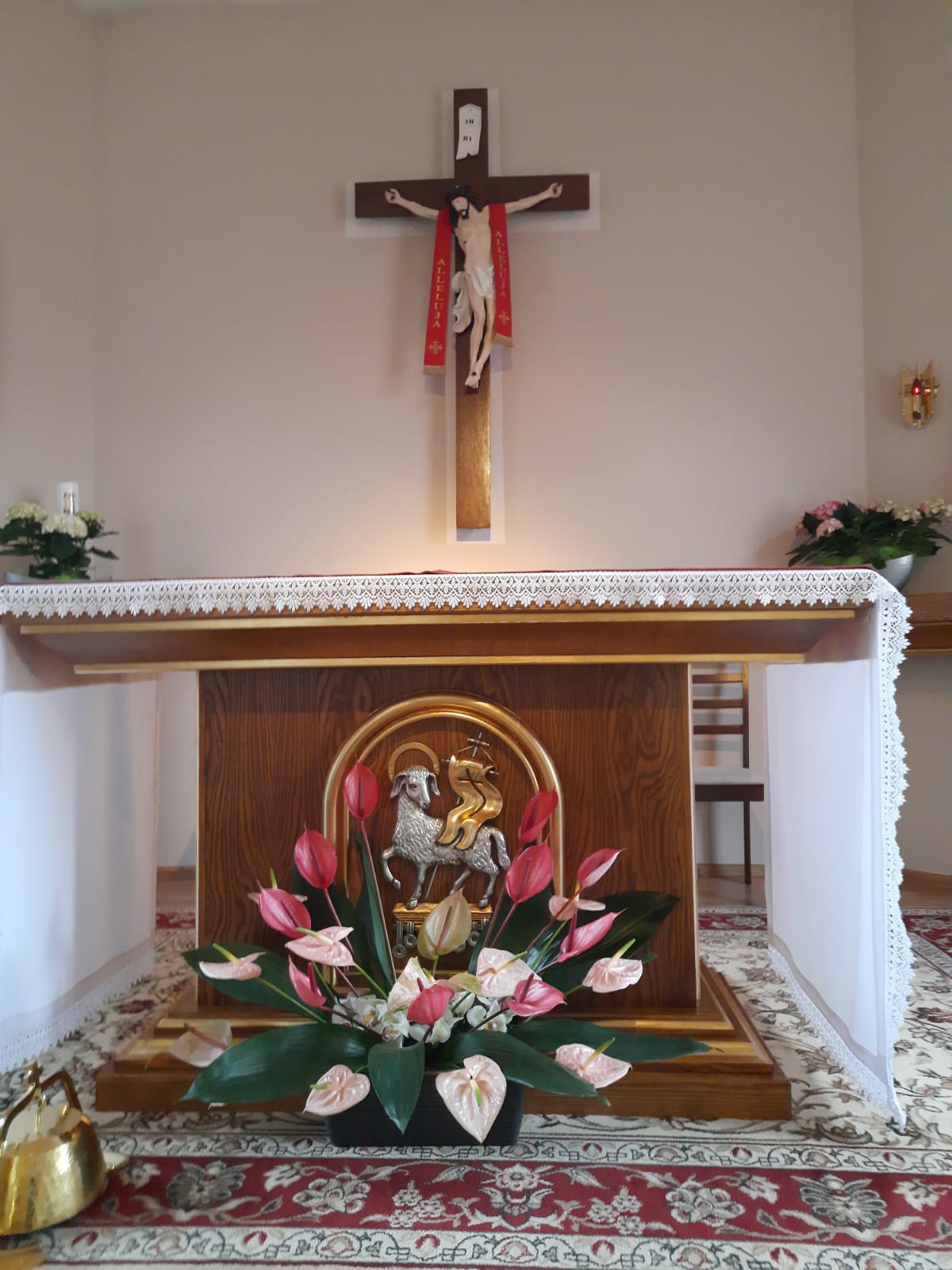 Zdjęcie przedstawia ołtarz naszej kaplicy, Przed ołtarzem stoi piękny bukiet kwiatów zza którego wyłania się wizerunek Baranka z proporcem, który symbolizuje Zmartwychwstałego Zbawiciela.