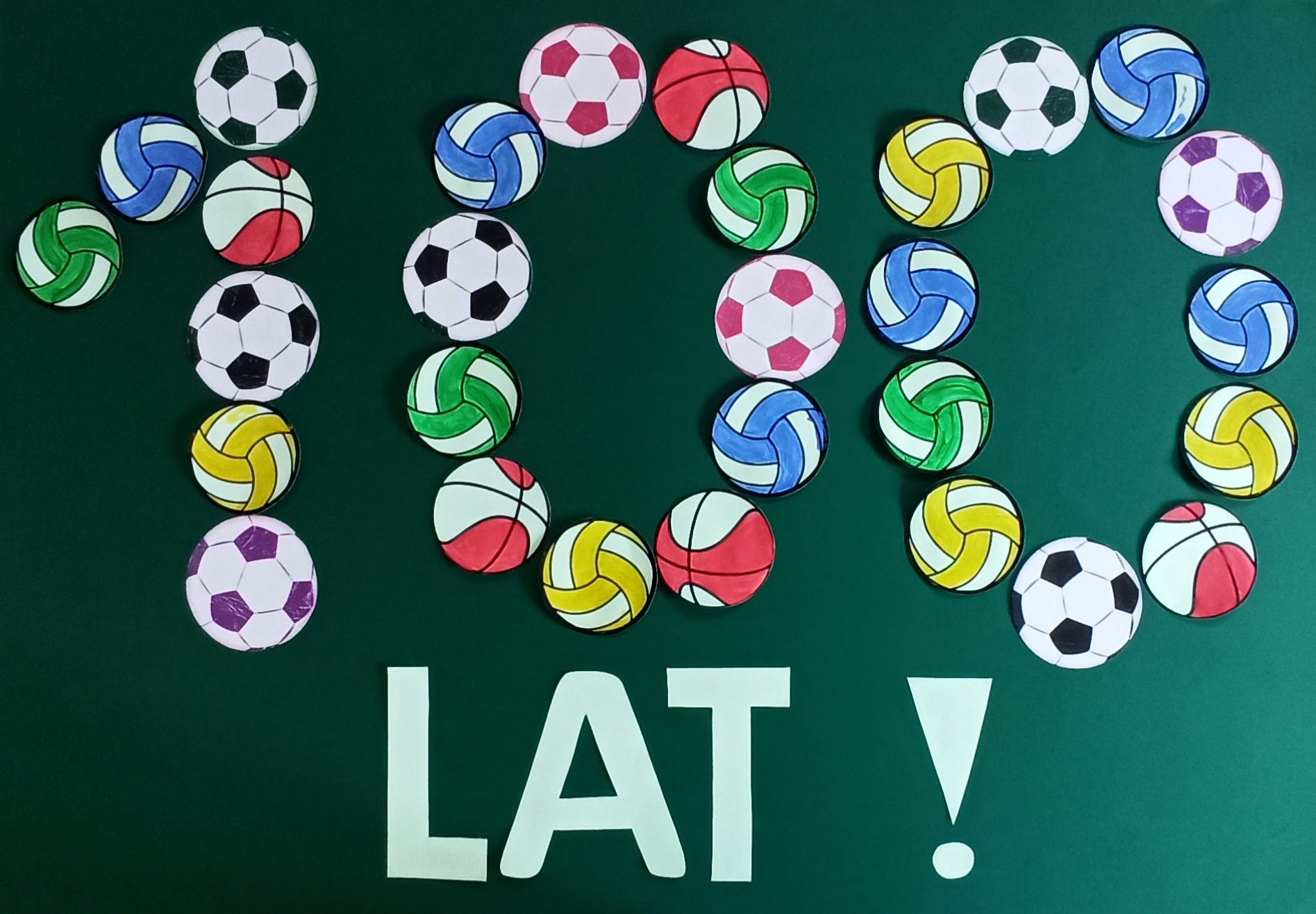 Napis "100 LAT" ułożony na dużym arkuszu zielonego brystolu z kolorowych pomalowanych farbami papierowych piłek do koszykówki, siatkówki i nogi.
