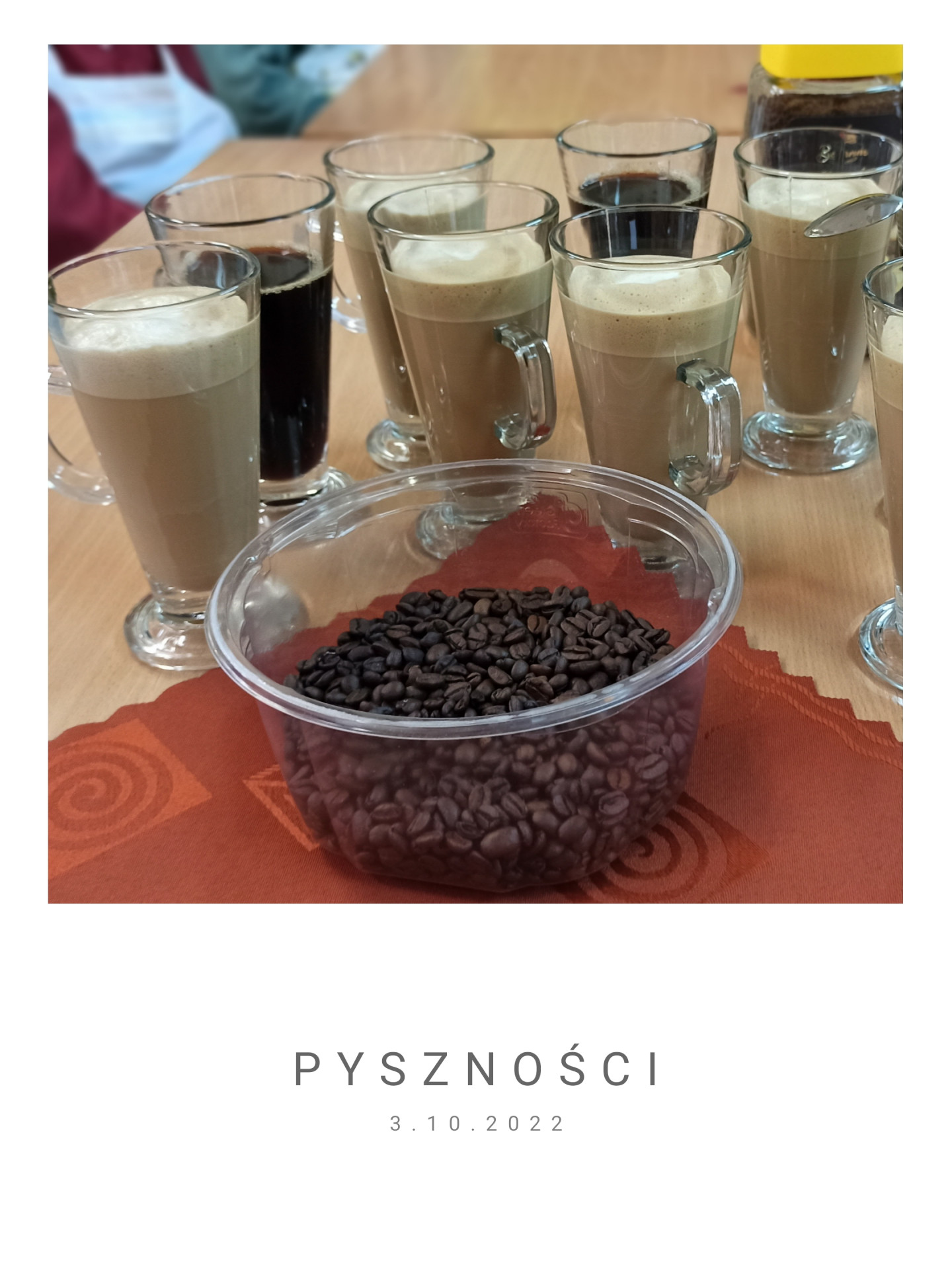 Obrazek ze zdjęciem przedstawiającym gotowe desery kawowe, kawy bez mleka oraz pojemnik z ziarenkami kawy na brązowej materiałowej serwetce. Na dole napis: „PYSZNOŚCI 3.10.2022”.