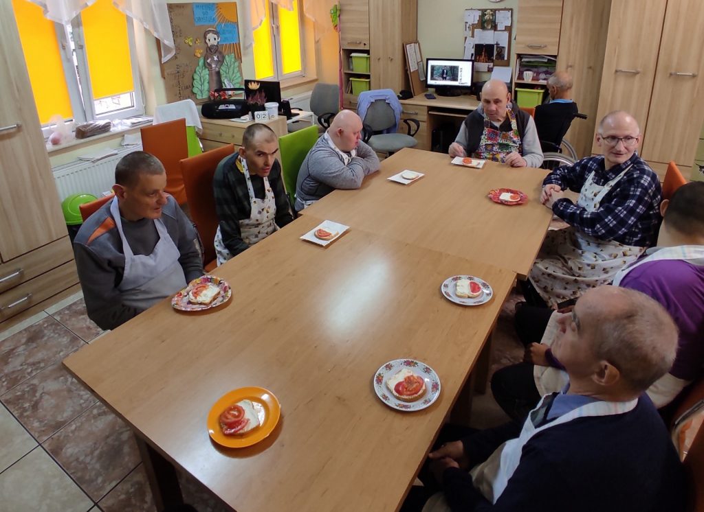 Grupa mieszkańców z oddziału drugiego siedzi w sali podczas zajęć kulinarnych. Przed każdym z mieszkańców jest talerzyk ze zrobioną kanapką. 