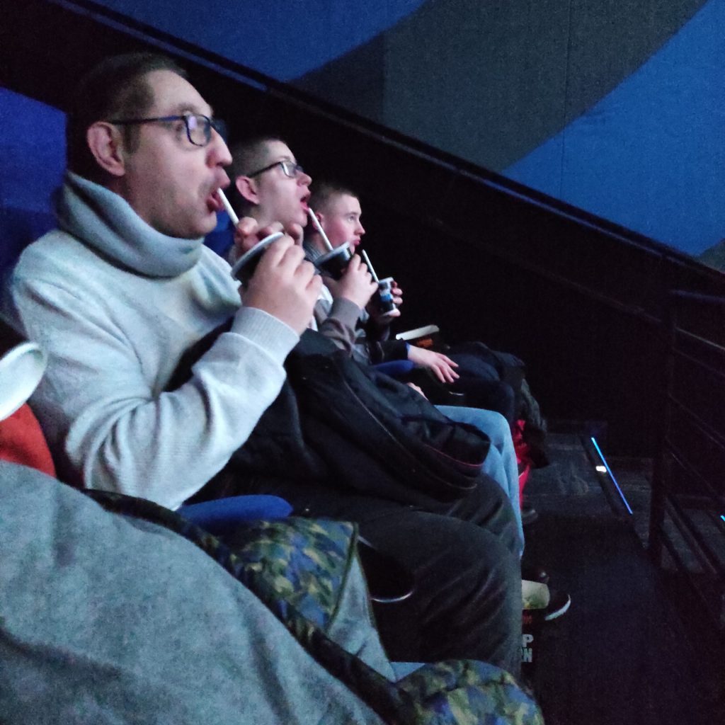 Mieszkańcy podczas oglądania filmu. Skupieni siedzą w fotelu na sali kinowej pijąc coca colę przez słomkę