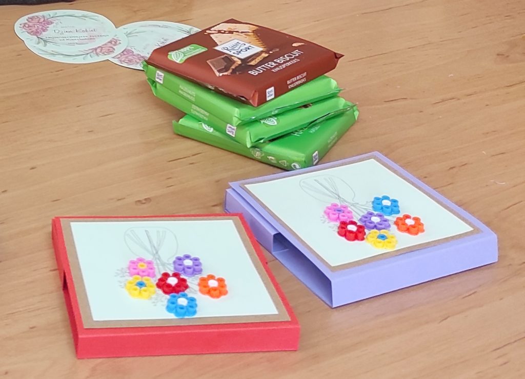 Gotowe pudełeczka na czekolady wykonane z kolorowego brystolu. Wieko przystrojone bukiecikiem z kolorowych kwiatków wykonanych z koralików do prasowania. Obok na stole leżą czekolady i stemple do spakowania.
